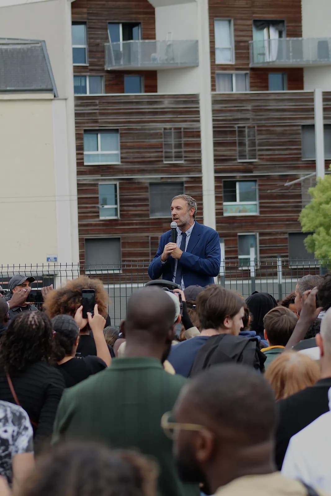 Un homme en costume bleu parle au micro devant une foule dense, au pied d’immeubles.