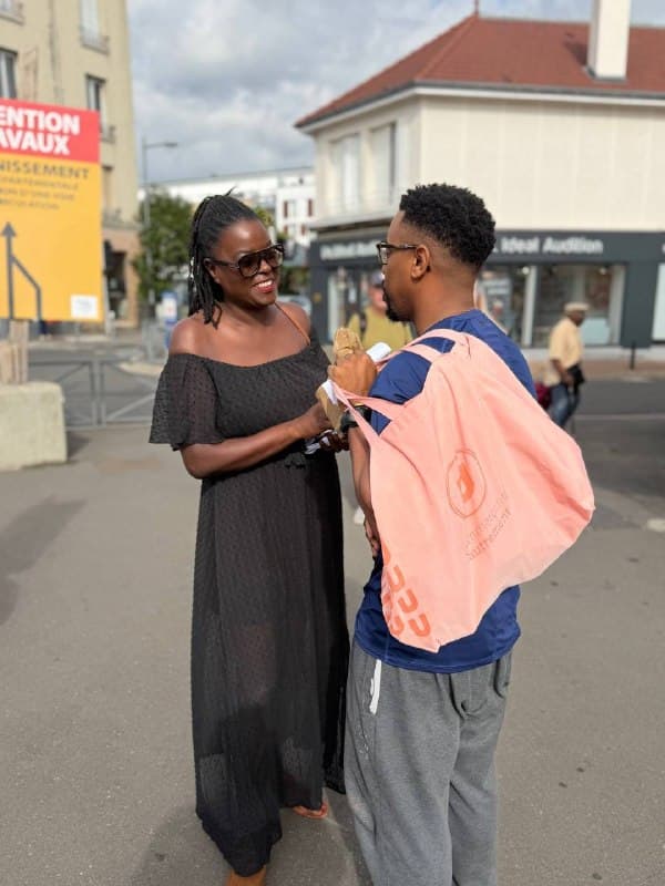 Une militante tend un tract à un passant chargé d’un sac, sur une place.