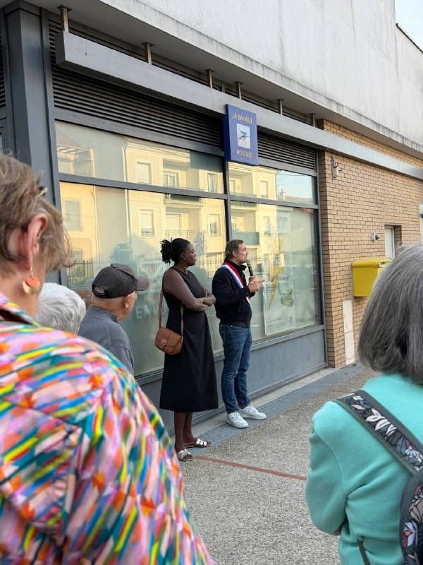 Le même homme au micro et la femme à ses côtés devant la vitrine, vus entre les habitants rassemblés.