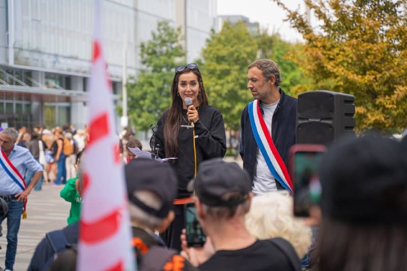 Une oratrice au micro devant le public, Thomas Portes en écharpe tricolore à ses côtés.