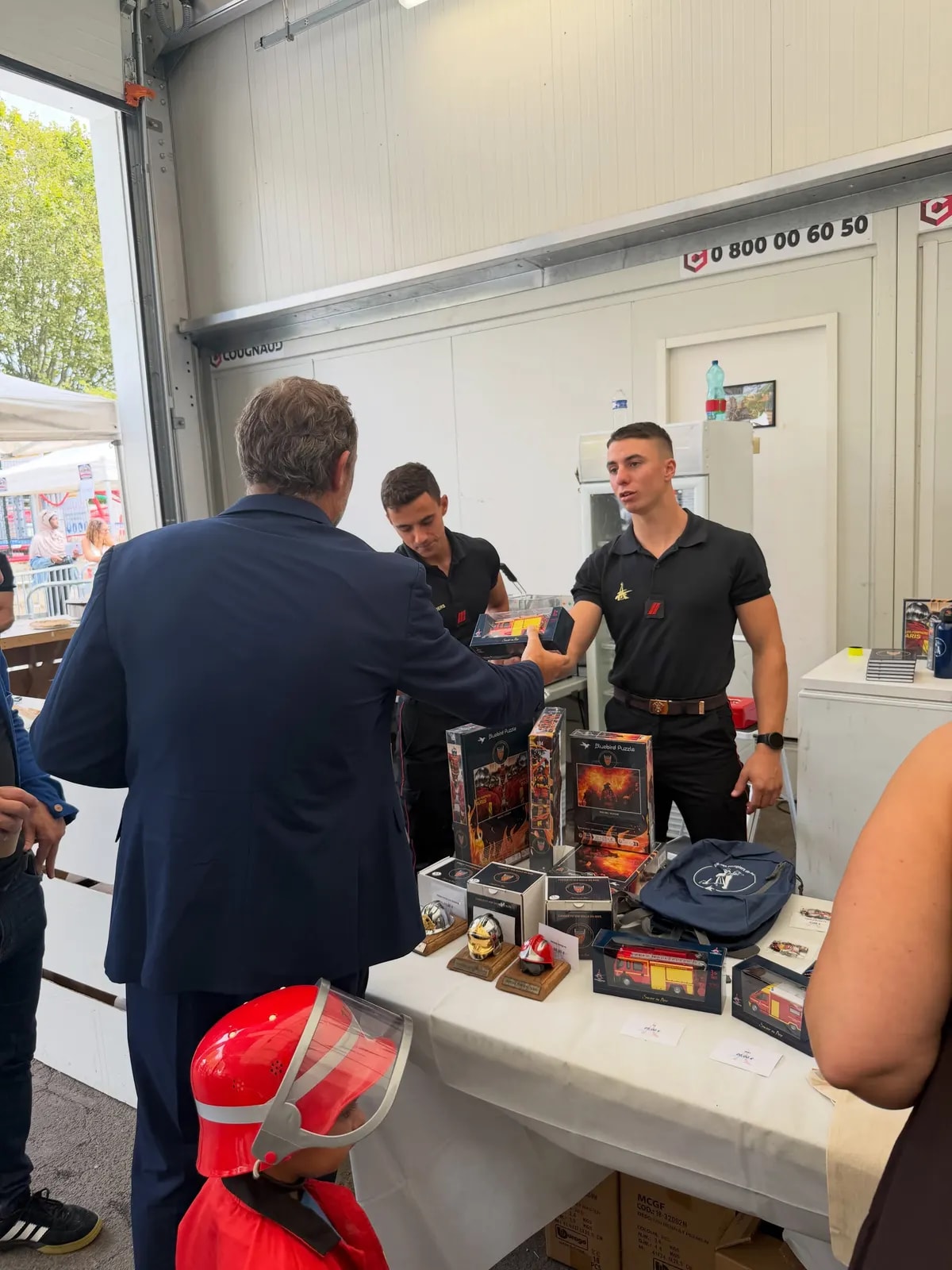 Un homme en costume s’entretient avec deux sapeurs-pompiers derrière la table d’un stand garnie de calendriers et d’objets.