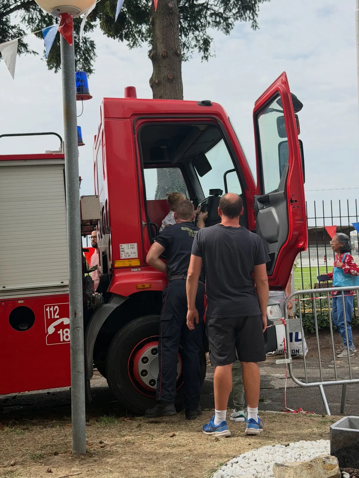 Un camion de pompiers rouge, portière ouverte, devant lequel des visiteurs attendent sous des guirlandes de fanions.