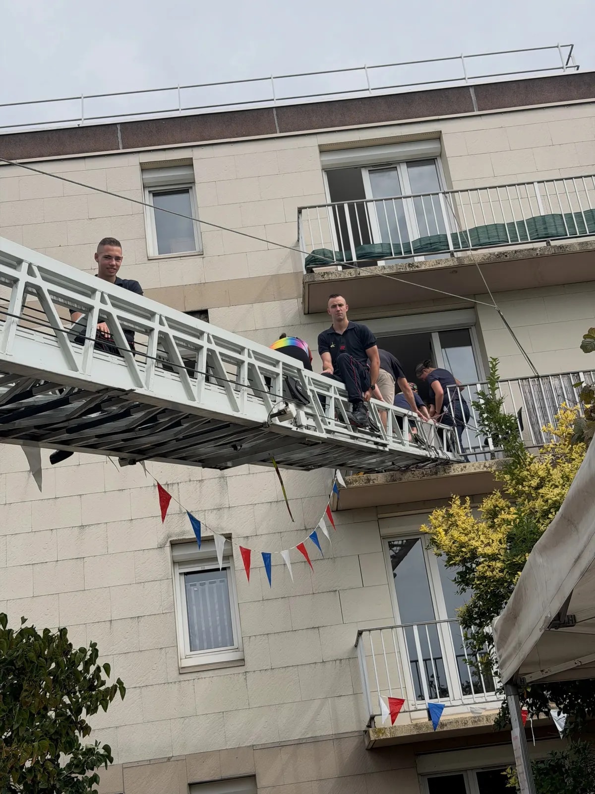 Une grande échelle déployée le long d’une façade d’immeuble, avec des pompiers et des visiteurs sur les échelons.