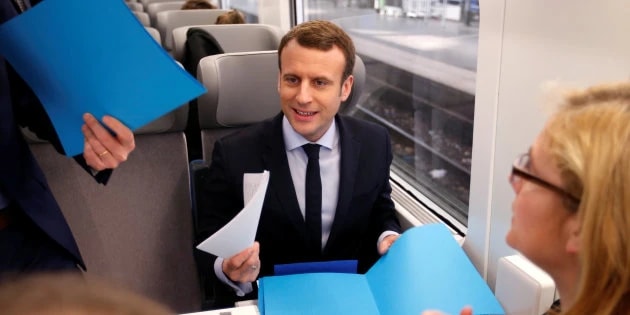 Homme souriant en costume assis dans un train, tenant des feuilles, face à une femme et à des chemises cartonnées bleues