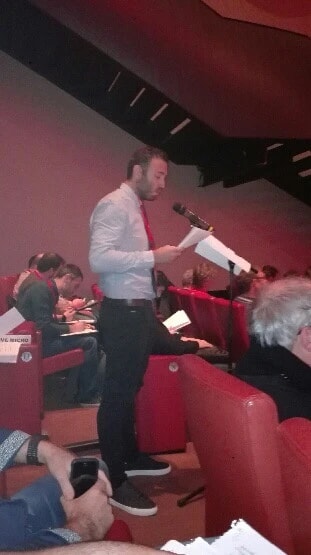 Un homme debout dans les gradins d’une salle de congrès prend la parole au micro, des feuilles à la main.