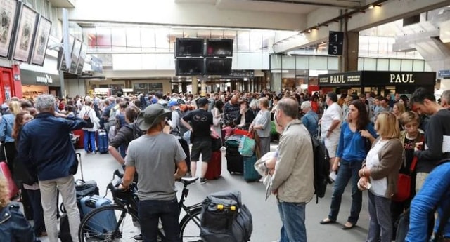 Hall de gare bondé de voyageurs avec leurs bagages devant les commerces.