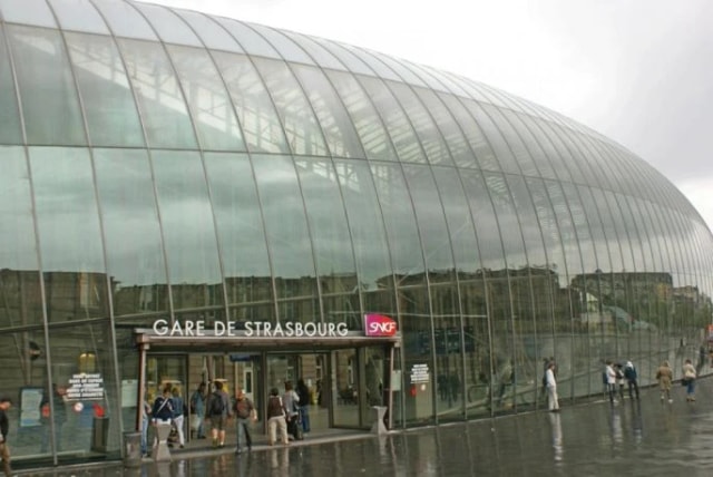 Façade de verre de la gare de Strasbourg un jour de pluie, quelques passants devant l’entrée.