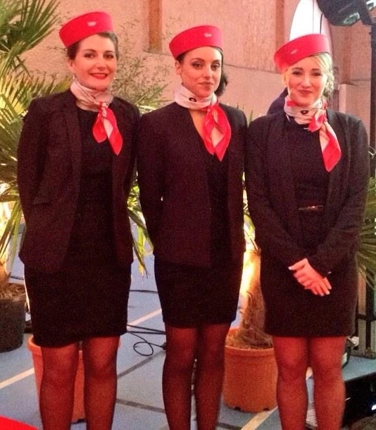 Trois hôtesses en tailleur sombre, calot et foulard rouges posent côte à côte lors d’une réception.