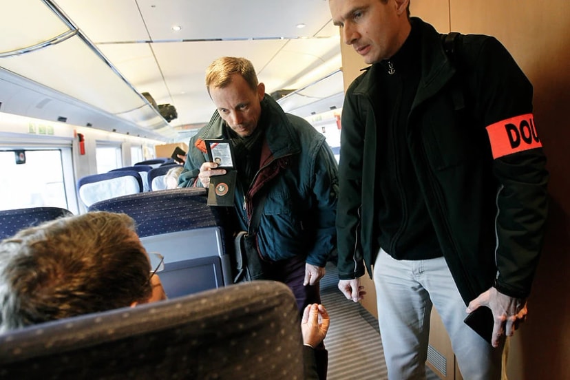 Dans une voiture de train, un homme montre une carte à un voyageur assis, à côté d’un homme au brassard orange « DOU ».
