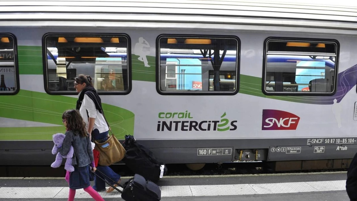 Une adulte et une enfant marchent avec leurs bagages le long d’une voiture Corail Intercités à quai.
