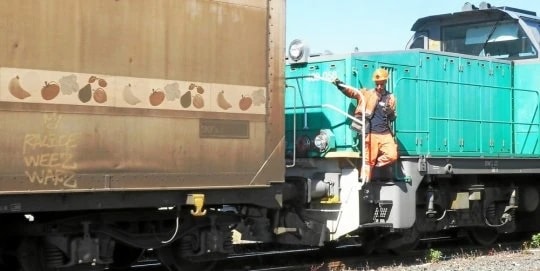 Wagon orné de fruits peints attelé à une locomotive verte, un agent en tenue orange sur le marchepied.