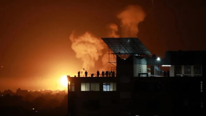 De nuit, une explosion illumine l’horizon d’orange et dégage d’épaisses fumées ; des silhouettes observent depuis un toit.