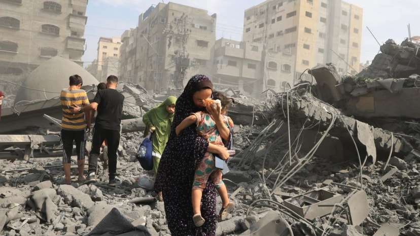Une femme porte un enfant dans ses bras au milieu des décombres d’immeubles détruits, d’autres personnes marchent derrière.