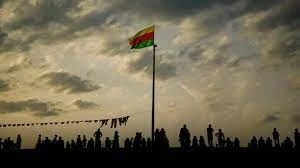 Drapeau vert, jaune et rouge flottant au sommet d’un mât, silhouettes d’une foule à contre-jour au crépuscule.