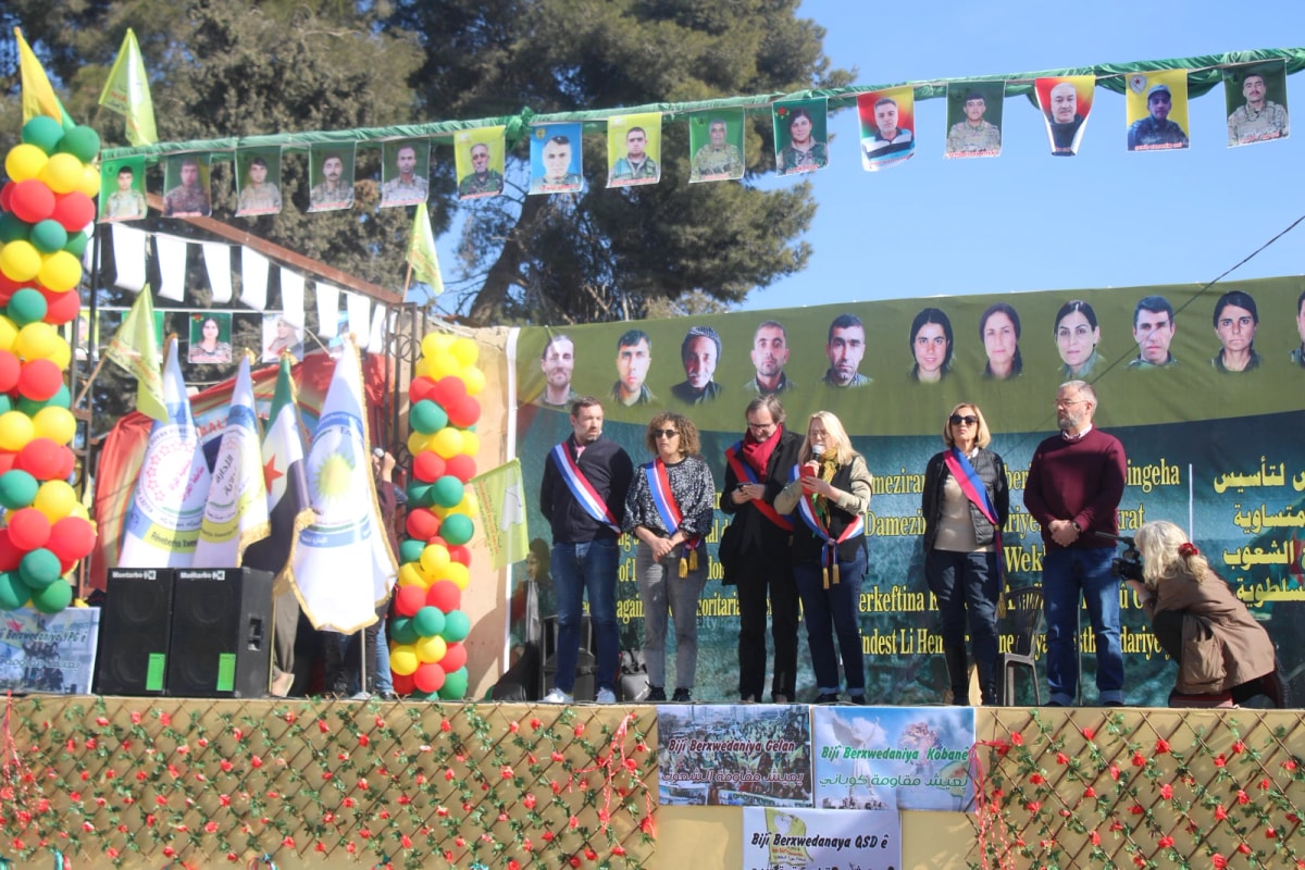 Estrade décorée de ballons devant une foule, plusieurs personnes en écharpe tricolore alignées sous un mur de portraits.