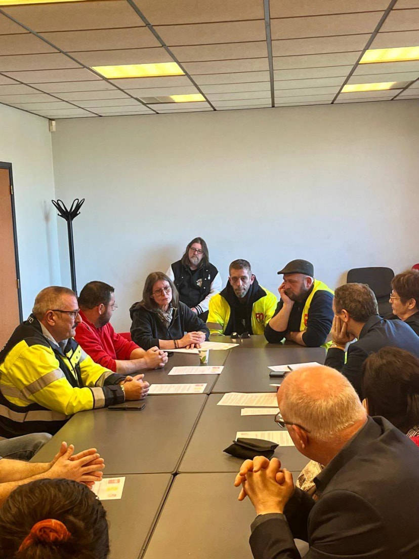 Réunion autour d’une grande table de salle de réunion, plusieurs participants en gilet de haute visibilité.