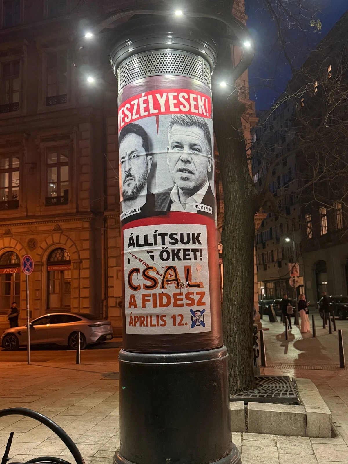 Colonne d’affichage couverte d’affiches politiques en hongrois, dans une rue le soir.