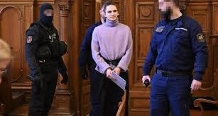 Une jeune femme en pull entourée d’agents en uniforme, l’un cagoulé, dans une salle boisée, le visage d’un agent flouté.