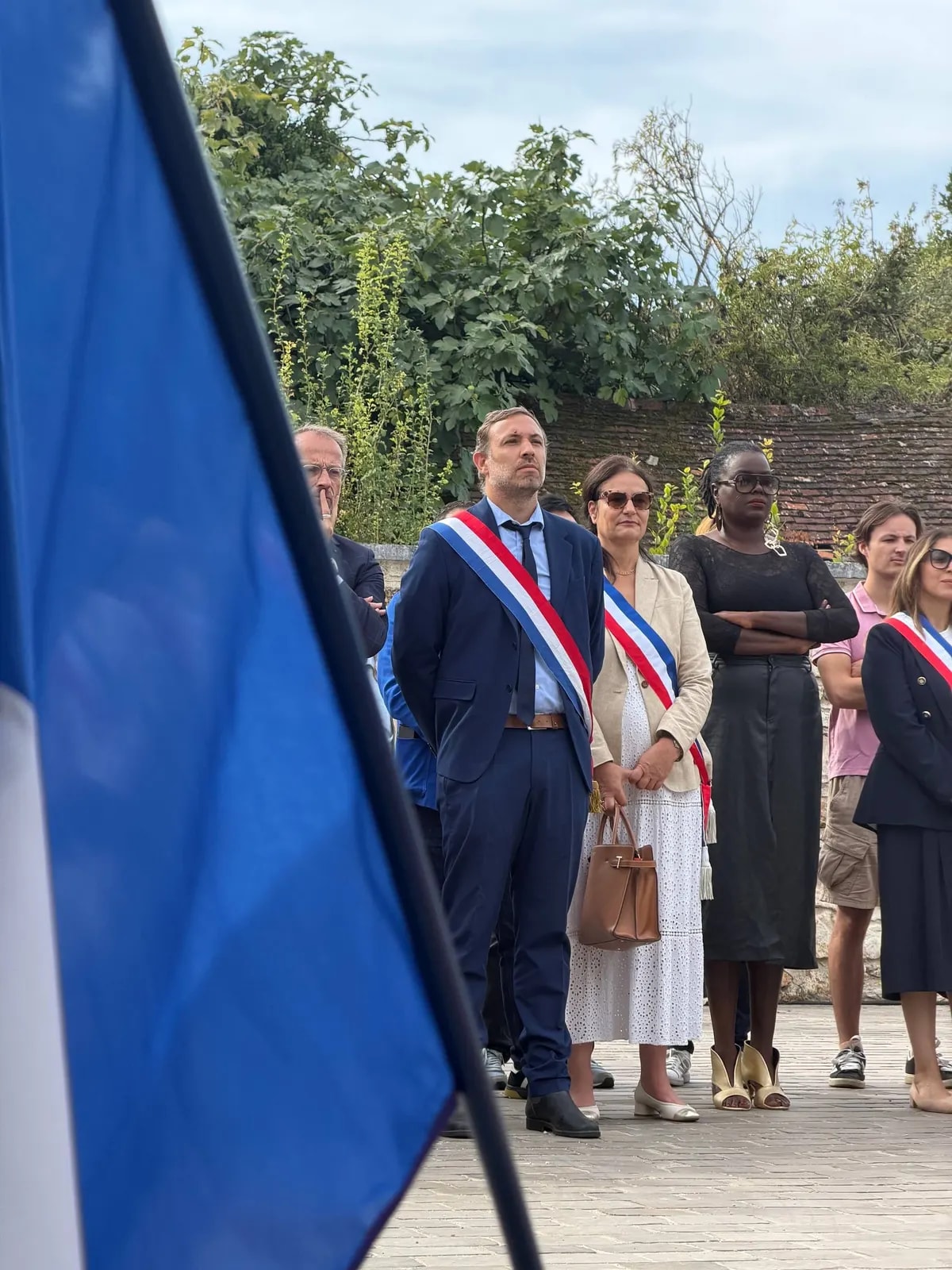 Des élus et participants devant un mur, un drapeau bleu au premier plan ; deux d’entre eux portent l’écharpe tricolore.