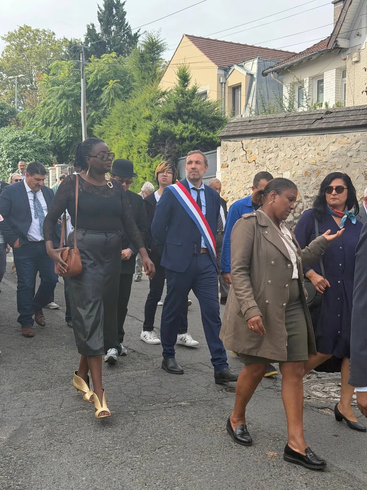 Un cortège remonte une rue résidentielle, précédé d’un élu portant l’écharpe tricolore.