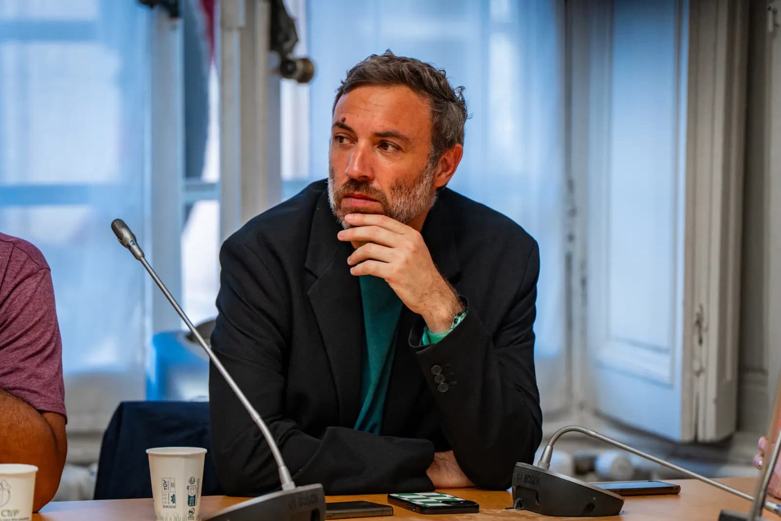 Un homme en veste sombre écoute, assis derrière un micro de table, dans une salle de réunion.